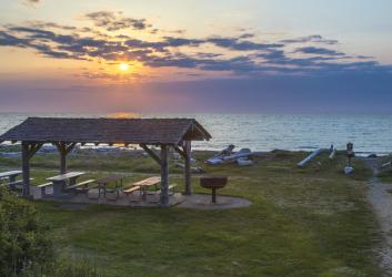 A wooden picnic shelter with picnic tables, surrounded by green grass, sits near the beach with driftwood nearby. The waters of the Strait of Juan de Fuca with a blue, yellow and pink sky as the sun sets behind the shelter.