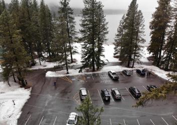 Cleanly plowed paved parking lot with marked parking spots bordered by pines and snowy ground next to a lake. Several trucks are parked in the lot.