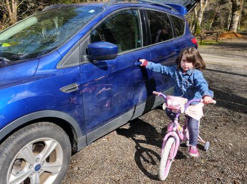 A little girl in a blue jacket on a pink bike with no helmet on grabs onto a blue car.