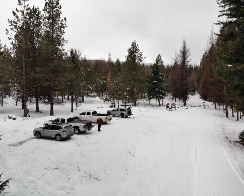 An unplowed parking area with compacted thin snow at a trailhead, next to a groomed road, surrounded by snow-dusted trees.