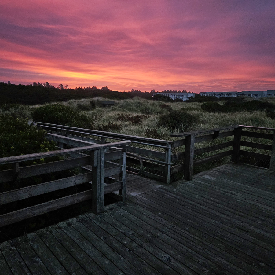 A pink and yellow sunrise cast over the sky while standing on an observation platform looking over dune grass and condos beachfront condos in the distance.
