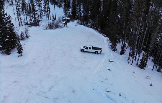 Overhead view of a parking area covered with compacted snow. A pit toilet hut lies on the edge.
