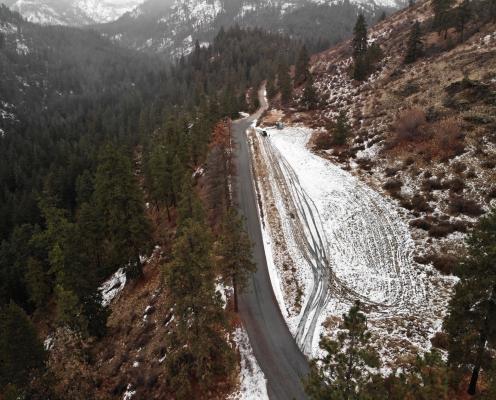 Overhead view of a long parking area covered by light snow running along a plowed highway, on a mountainside covered by pine trees. A porta-potty lies at one end.
