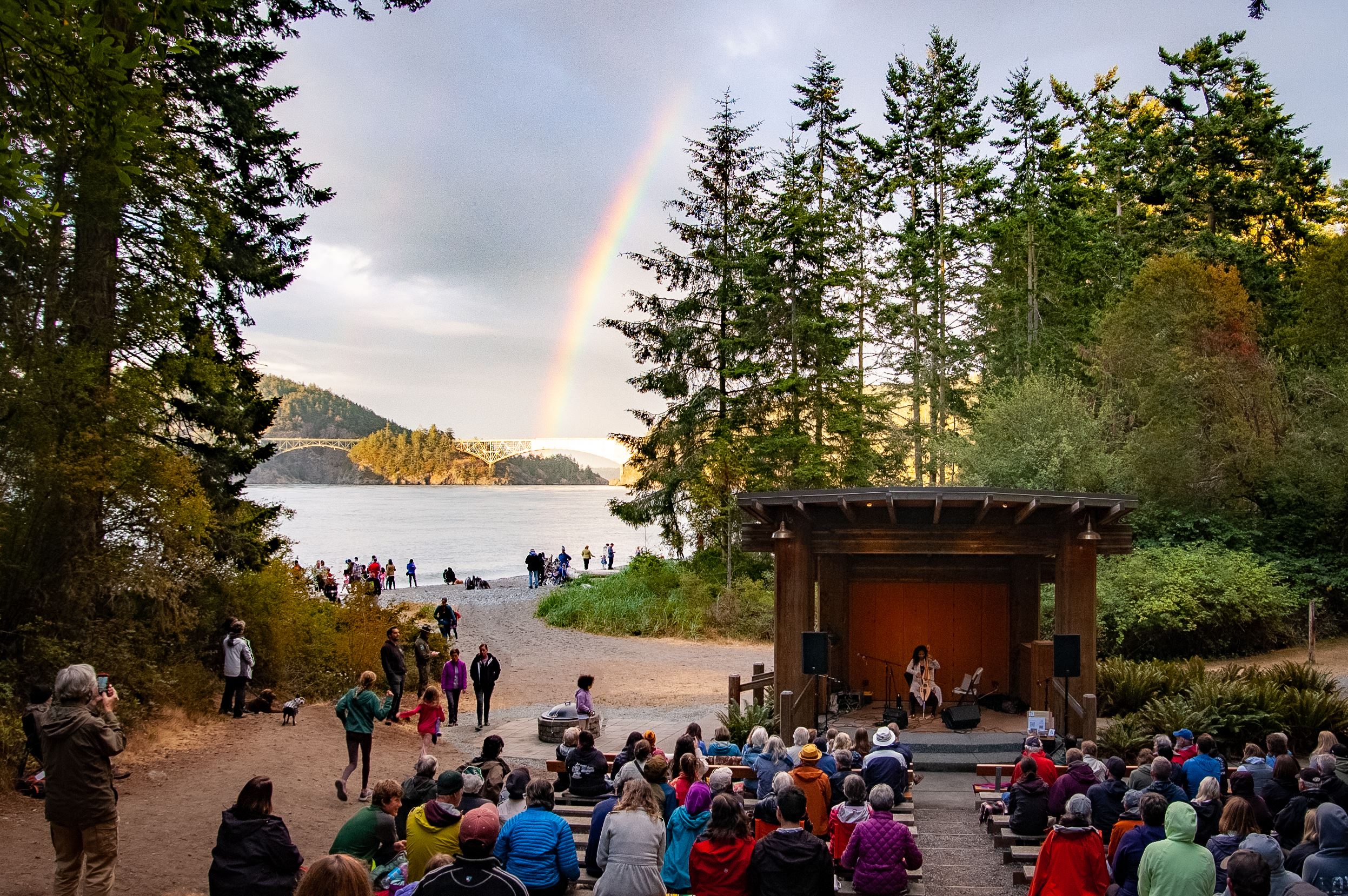 A band playing in an amphitheater at Deception Pass State Park with an audience watching and water, trees and a rainbow behind.