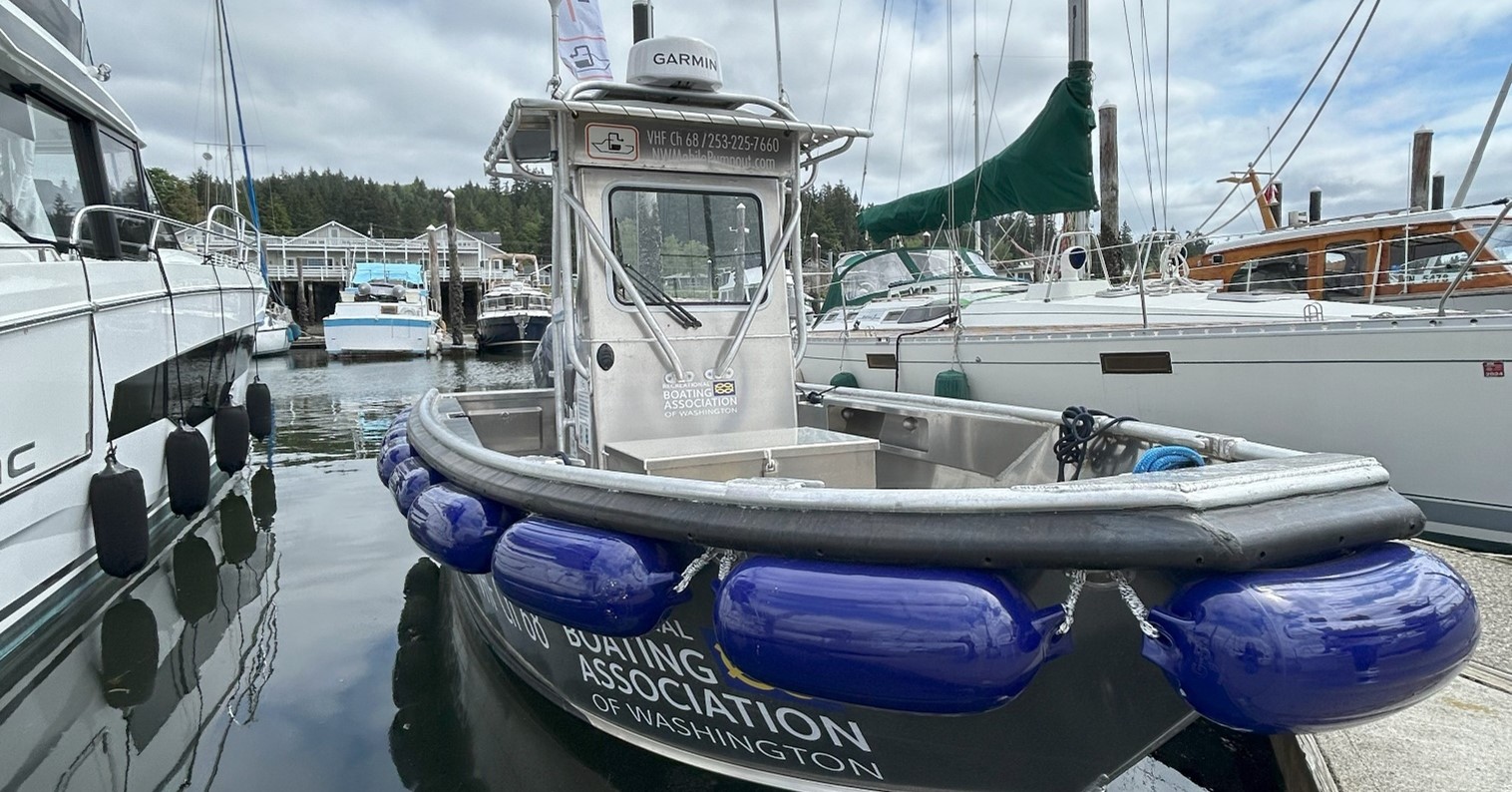 A boat is in the water at a marina. There are blue buoys secured around its bow. The water is still.