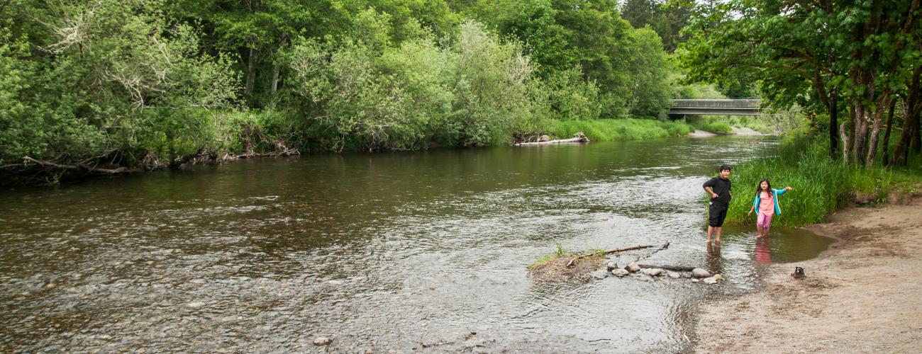 Two youths enjoying the river shoreline. There are rocks visible by the water, trees with green leaves visible on both sides, and in the distance what looks to be a bridge over the water. 