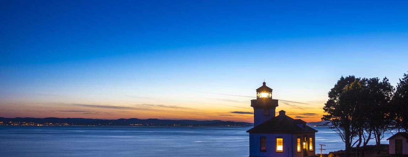 An illuminated lighthouse sits on a rocky cliff overlooking water as the sun sets behind and the sky grows dark. A star shines bright in the sky.