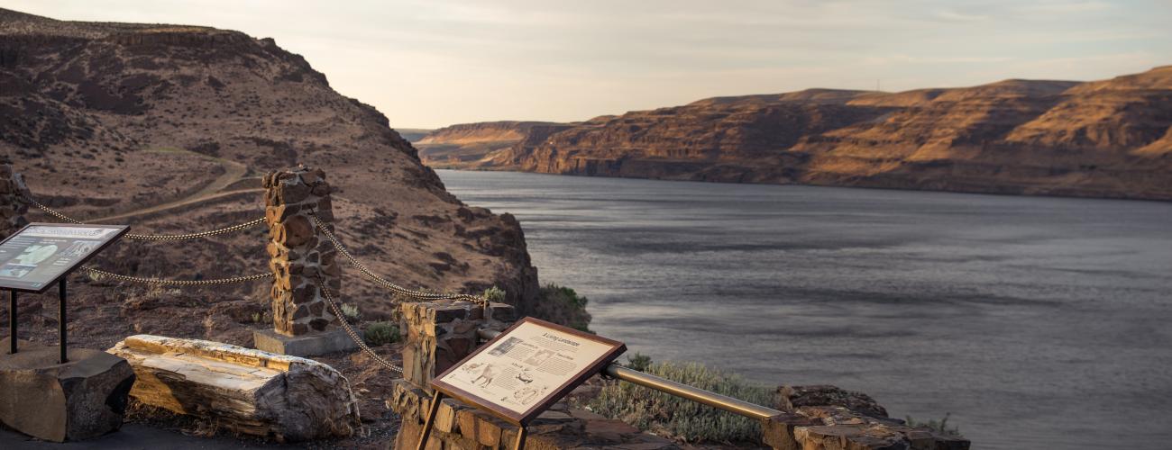 Interpretive panels sit in front of a rock wall with a chain rope with the Columbia River in the background.