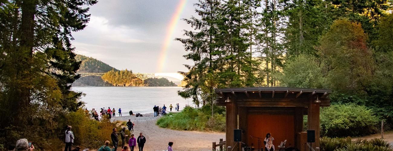 A band playing in an amphitheater at Deception Pass State Park with an audience watching and water, trees and a rainbow behind. 