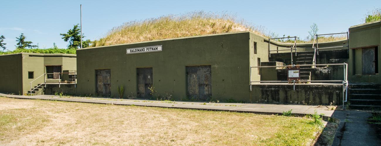 A cement battery building sits with brown grass in front and on top. Doors are boarded shut with stair ways throughout the structure.