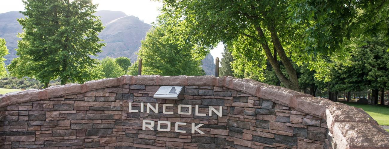 Rock wall structure with 'Lincoln Rock' with tall, leafy trees surrounding the structure. In the distance, a hillside can be seen.