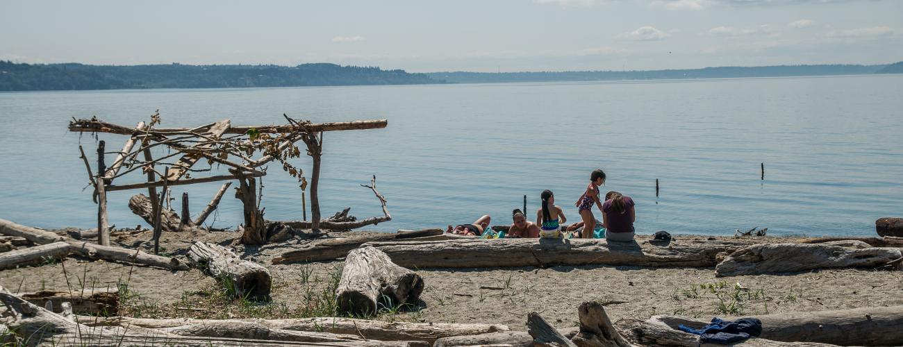 a group of kids sit on a log on the beach next to a driftwood arch