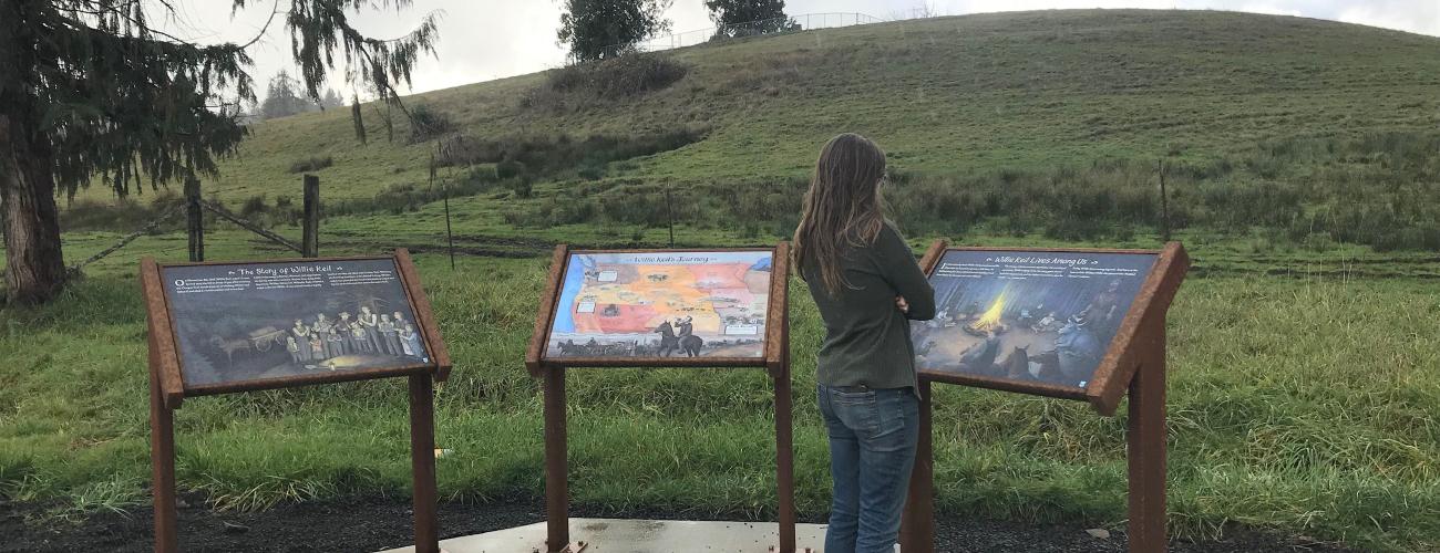 A person with long hair and wearing a T shirt and jeans looks at one of three interpretive panels set in a semi-circle on a concrete pad. A grassy hill with a few sparse evergreen trees on it rises under a cloudy sky in the background.