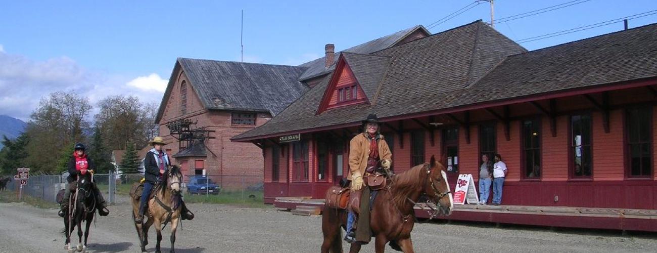 Three horse riders on brown, tan and black horses walk on a dirt road in front of an old trail stations painted orange with red trim. Two people wearing jeans and tshirts lean against the building watching the horse riders go by. 