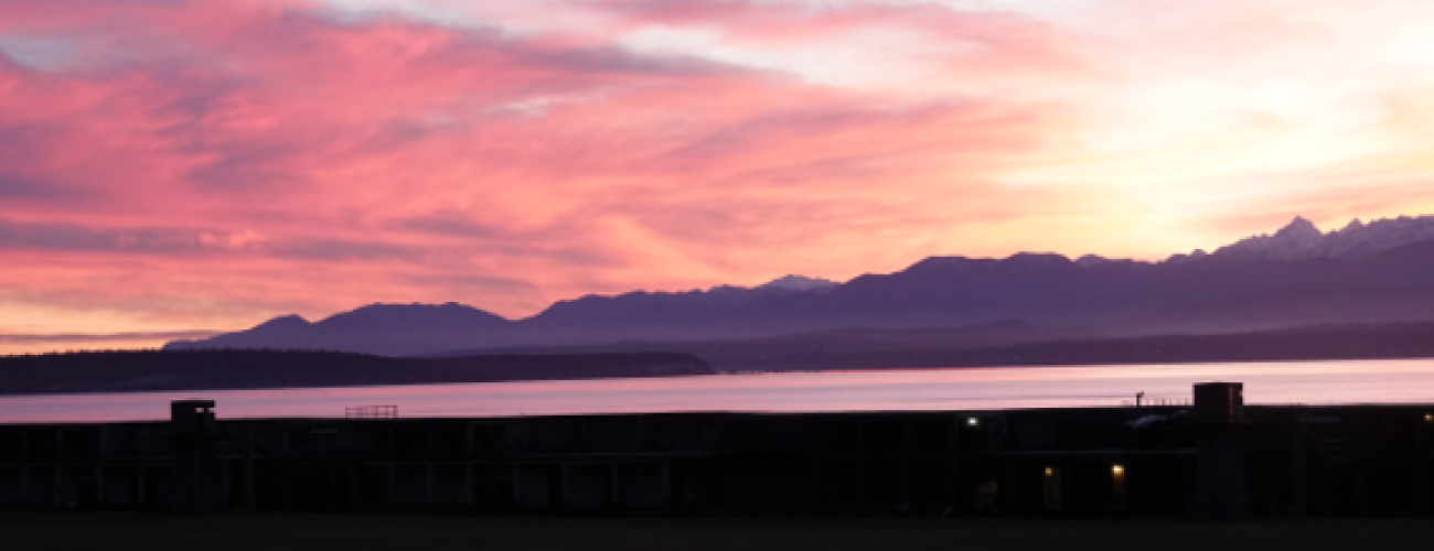 Sunset from Fort Casey: Olympic Mountains backlit by sun setting on left side of image, clouds changing from golden yellow to salmon orange to pink edged in purple. The fort is a black silhouette with just the tops of the battery commanders' towers visible in the pinkish waters of the Salish Sea.
