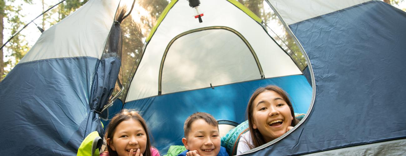 Three children poke their heads out of a tent. They are smiling.