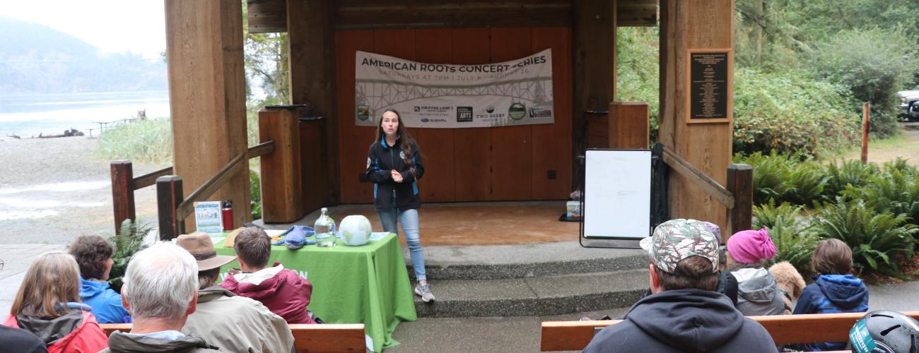 AmeriCorps staff stands in amphitheater facing audience as she prepares to talk to park visitors. Visitors sit in wooden benches watching staff. 