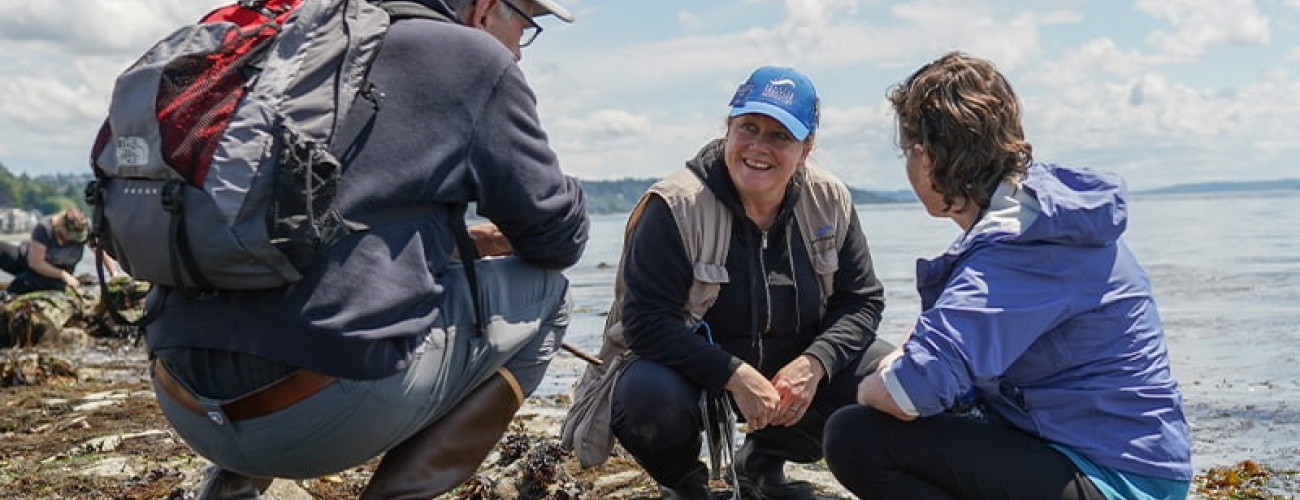 Three people squatting on the beach engaged in a discussion. In the middle is the Seattle Aquarium Beach Naturalist who is a feminine appearing person in a blue Seattle aquarium hat wearing a khaki colored vest and speaking to a masculine looking person to their left in a grey sweatshirt and jeans with a red and grey backpack. To the right of the beach naturalist is a feminine appearing person with brown hair and a purple jacket with black plants.