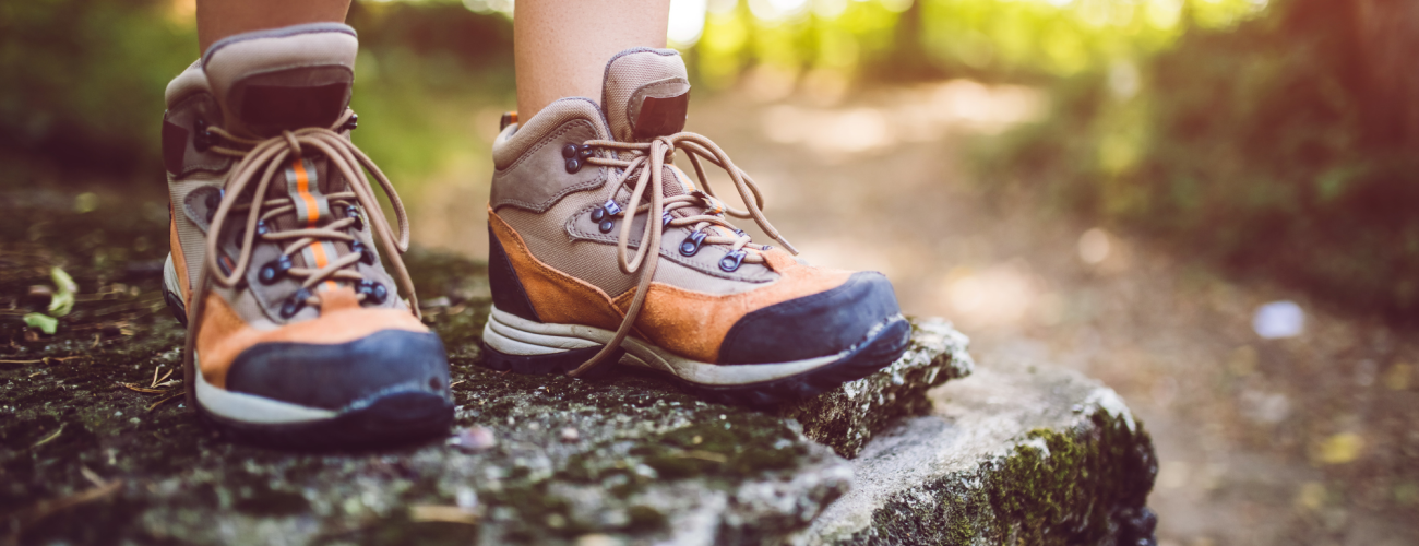 Children's brown, orange, and black hiking boots in focus with a trail out of focus in the background. Child is standing on a rock that is semi-in focus and only the child's shins down are visible. Child isn't wearing socks. 