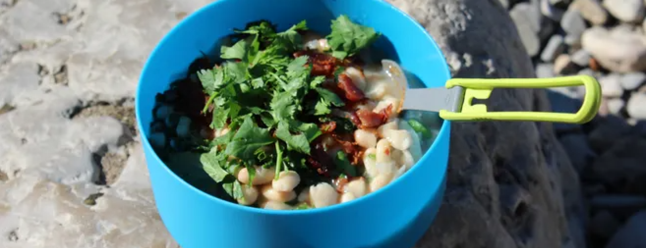 Blue bowl on a rock filled with beans, cilantro and bacon. A foldable spork is pressed into the mixture