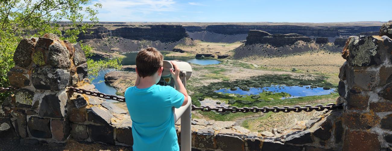 A boy in a blue shirt and shorts using the provided binoculars to lookout over the ice age flood rock formations at Sun-Lakes Dry Falls State Park. This view can be found at the parking lot of the Dry Falls Visitor Center.