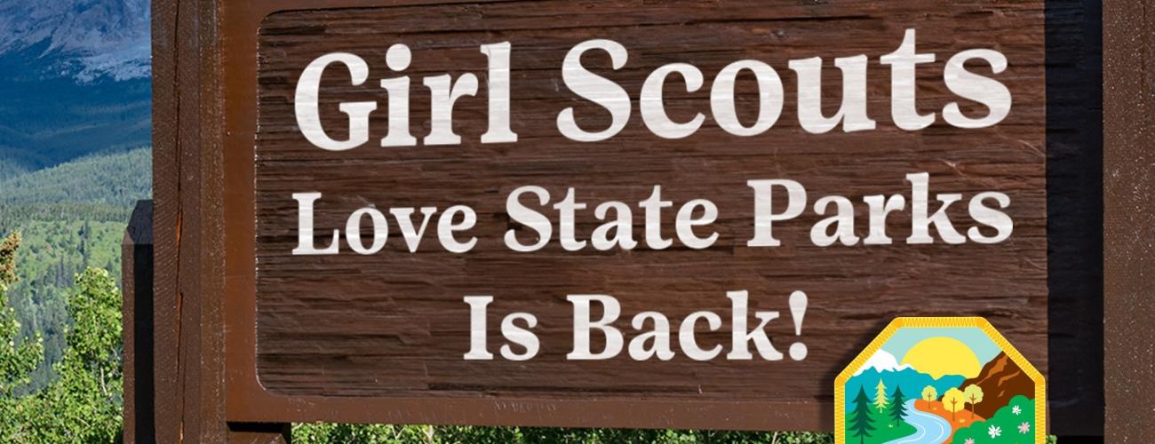 Set along the roadside, with green bushes, a mountain and blue sky are set behind a brown wooden sign that reads "Girl Scouts love State Parks is back!". A smaller sign in front of the wooden sign reads 'Girl Scouts love State Parks' with a river and mountain scenery. 