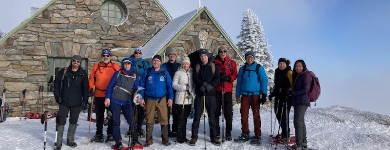 A group of snowshoers in colorful winter clothes stands on a field of fluffy snow in front of a snow covered stone cabin. Pale clouds rise into a clear blue sky behind them.