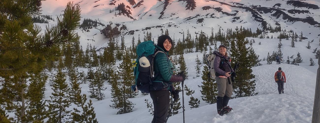 Two women snowshoe at sunrise below a snowy mountain.