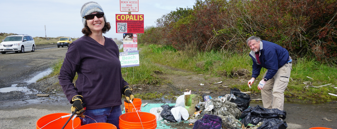 Woman and man posing for a photo at the beach with buckets in hand in front a pile of trash