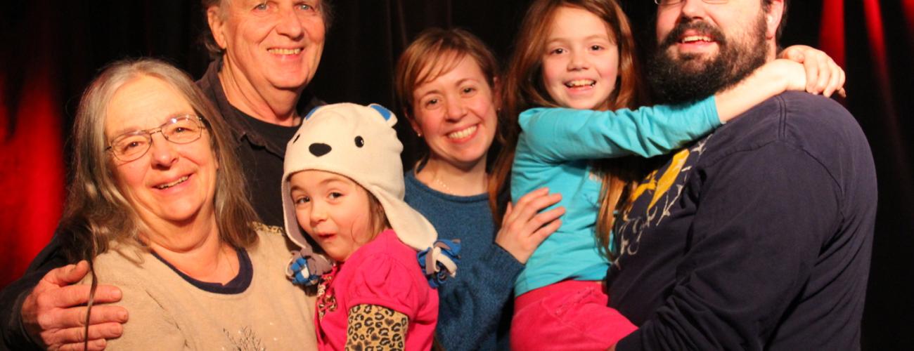 A multi-generation musicians standing and two of them holding a child in their arms, black background with red curtains on both sides