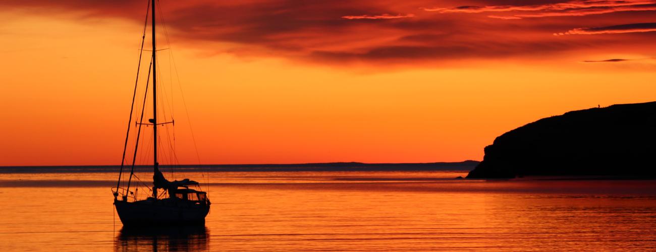 View of orange sunset looking over a saltwater bay. One sailboat and island in the background.