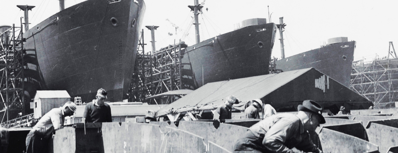 Black and white photo where six men are working on building a part of a ship. In the background, three ships further along in construction are visible.