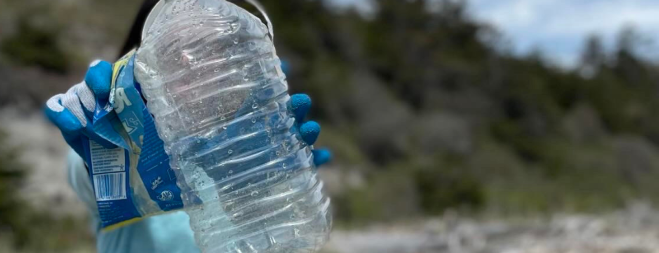 Person holding gallon water bottle and candy wrapper with blue gloves on and beach in background