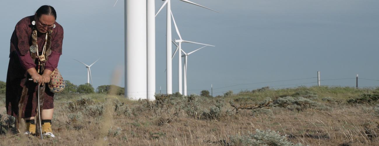 Native American putting a stick into the ground, wind turbines on the background
