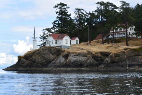 A picturesque red and white light station building next to a device that provides the light. It stands atop a rocky precipice with evergreen trees behind it. 