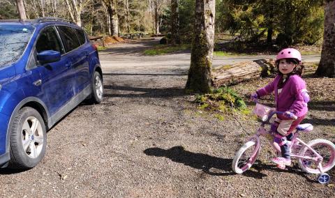 A little girl in pink on a pink bike with a pink helmet stops for a blue car.