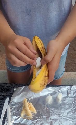 Person in a grey shirt and shorts sticks marshammaows into hollowed out banana