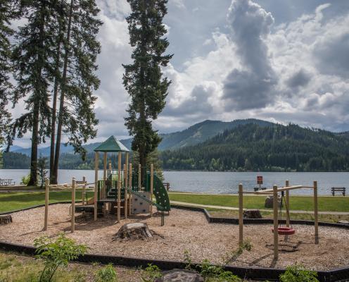 An outside child play structure with adjacent tire swing sitting on play bark. Located off to the side of the green grass behind the swimming area. Dark green evergreens across the lake and white clouds against a blue sky. 