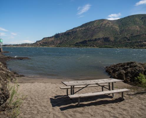 Sandy beach with picnic table next to shoreline lake cliffs in the distance and green boat guide marker light