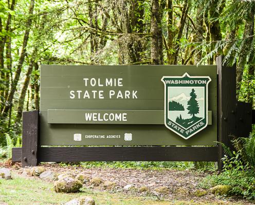 Tomie State Park welcome sign. Sign is greenish-brown with chocolate brown framing. Sign is wooden and surrounded by lush, green trees. 