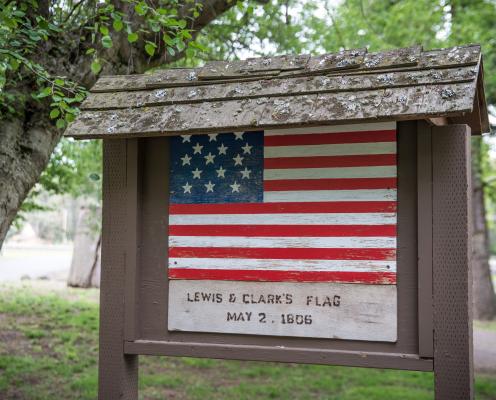 Brown roofed sign with a painted replica of Lewis and Clark's flag from 1806 with 15 stars on a blue background and 15 red and white stripes