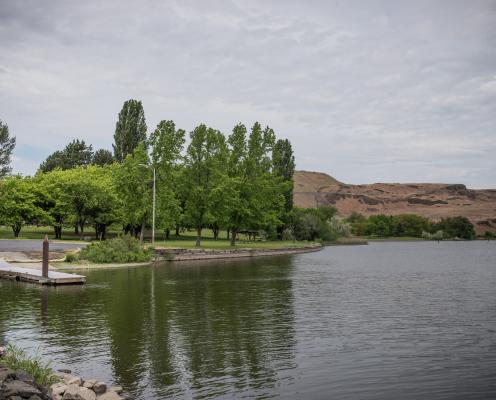 Green trees sit on a grassy lawn that goes to the water's edge. A boat launch sits on the left side of the lawn with a brown, semi-arid shrubsteppe hillside in the background.