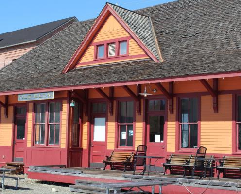 Old western style building painted orange with red trim with a sign on the left front reading, "Cle Elum". Wooden park benches sit on the front, open porch of the building. Picnic tables sit on the dirt in front of the building.