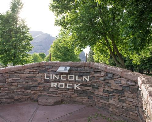 Rock wall structure with 'Lincoln Rock' with tall, leafy trees surrounding the structure. In the distance, a hillside can be seen.