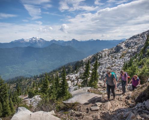 Three hikers hike the trail through dirt and rocks, surrounded by large boulders and evergreen trees scattered through the boulders. In the background is treed mountains with a large, snow capped mountain top with a blue cloudy sky overtop. 