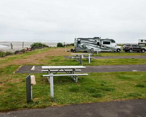 RV Camping parking spaces with picnic tables and small lawns right against the Pacific Ocean shore. A few RVs are parked in the spaces at the far end of the campground.