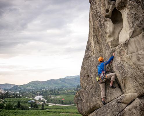 A person in a blue shirt and orange helmet scales a granite rockface using ropes. They look toward the distant verdant valley and mountain ridges under a steely gray cloudy sky.