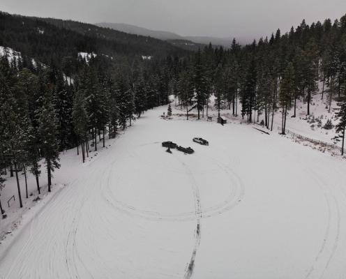A open snow-covered area with circular tire tracks surrounded by trees with snowy hills in the background. A pit toilet stands in a corner.