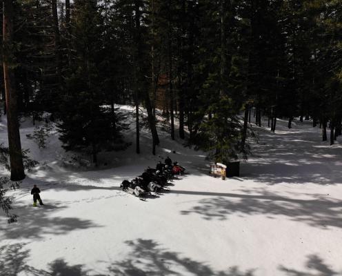 A shady forest road covered in deep snow leading to a parking area surrounded by trees. Five snowmobiles and their riders sit in the middle.