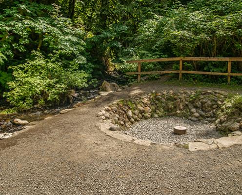 A circular pit surrounded by trees and a wooden fence. It is lined with stones and in the middle is a round stone with a hole in it. This is where the flame of Flaming Geyser was once constantly burning.